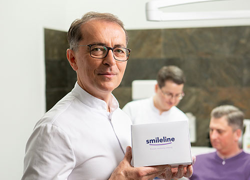 Profesor Fudalej trzymający białe pudełko z napisem Smileline w tle dwie inne osoby 1