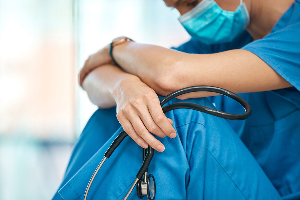 Osoba w niebieskim uniformie medycznym i w maseczce, trzymająca stetoskop, siedząca z podkulonymi nogami 1